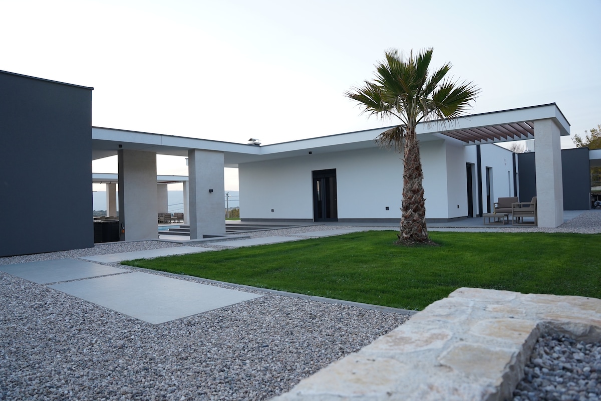 A modern residence is surrounded by a landscaped garden featuring a palm tree. The structure's exterior includes large windows and a combination of stone and smooth finishes. Paved walkways lead to various outdoor seating areas, providing a harmonious blend of nature and design.