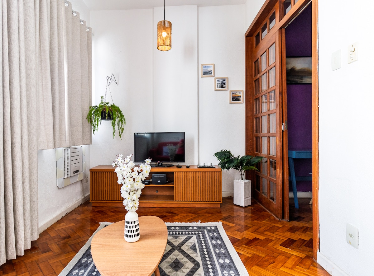 A living area is showcased with a television atop a wooden media unit, complemented by a decorative plant. Natural light fills the room, accentuating the wooden flooring and textured curtains. A geometric coffee table sits centrally, surrounded by a cozy atmosphere.
