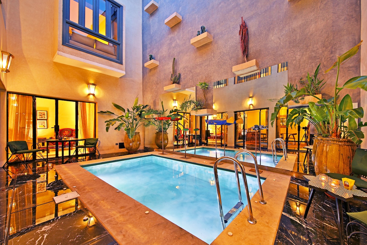 A serene swimming pool is framed by vibrant plants and natural stone walls, creating a relaxing ambiance. Large windows allow sunlight to illuminate the space. Comfortable seating areas surround the pool, providing ideal spots for relaxation and socializing.