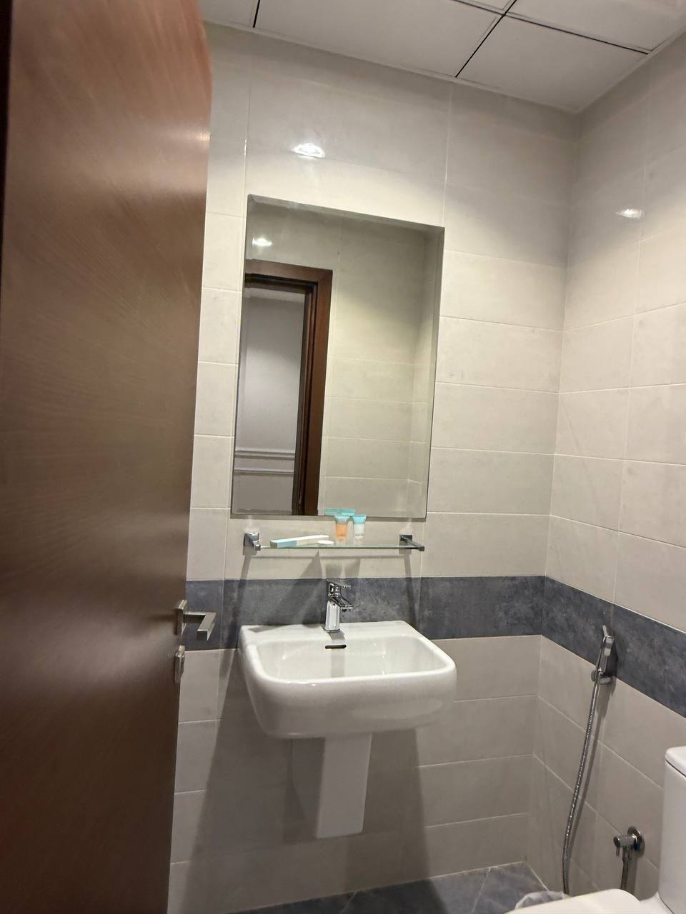 A compact bathroom features a modern sink with a square basin and a large mirror above. Light-colored tiles line the walls, creating a clean aesthetic. A towel rack is mounted nearby, and a showerhead is positioned in the corner.