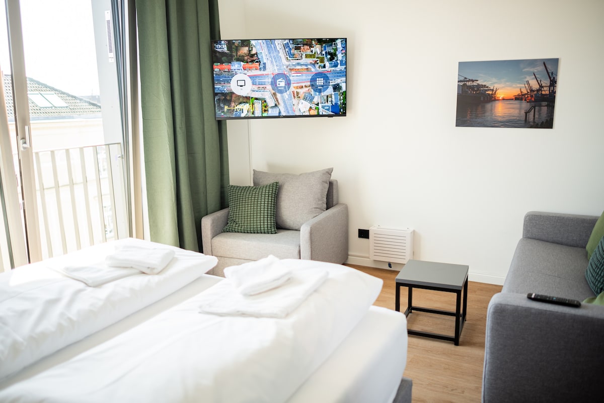 A comfortable sleeping area is furnished with a spacious bed layered with white linens and two rolled towels. Nearby, a light grey sofa and chair are positioned next to a small table. A large television is mounted on the wall, and a scenic photo is displayed.