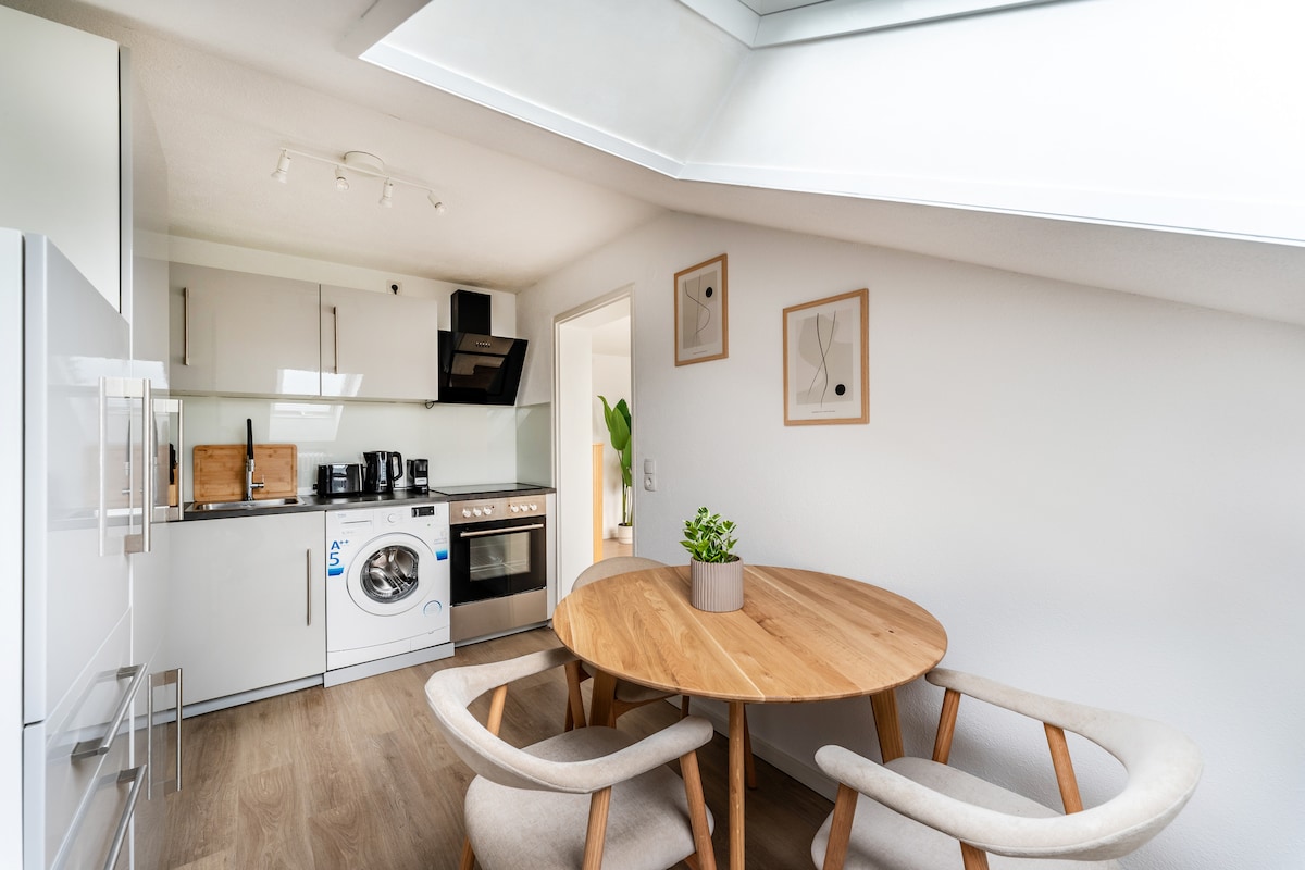 A modern kitchen is equipped with sleek white cabinetry and high-quality appliances. A round wooden dining table is accompanied by two light-colored chairs. The space is illuminated by natural light filtering through a skylight, with minimalist decor enhancing a clean aesthetic.