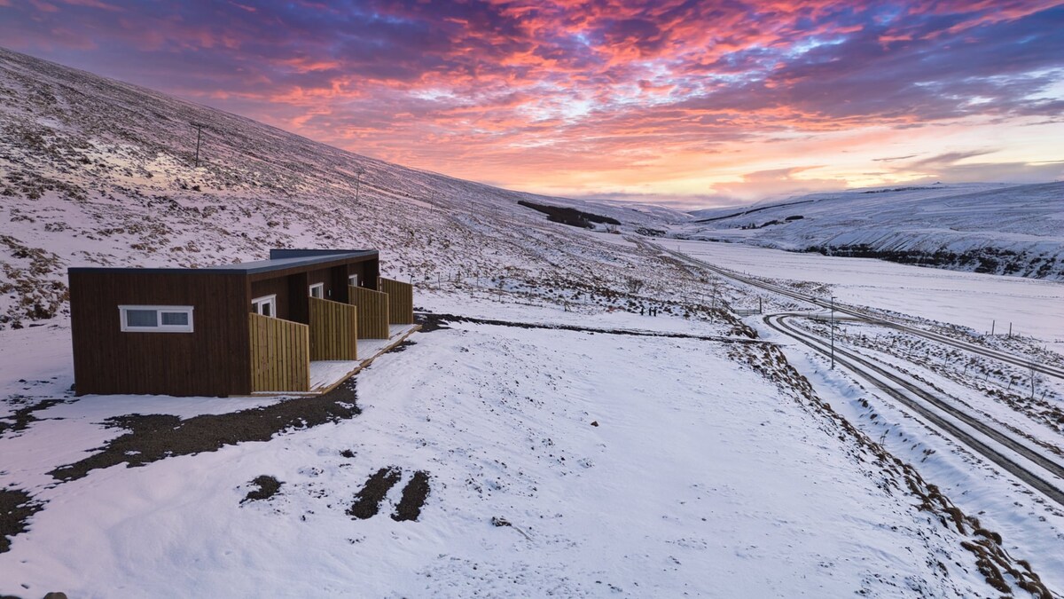 A modern cabin is positioned in a snow-covered landscape, with dramatic mountain scenery in the background. The sky displays a colorful sunset, casting soft light across the snowy ground. A winding road leads towards the cabin, emphasizing the serene setting.