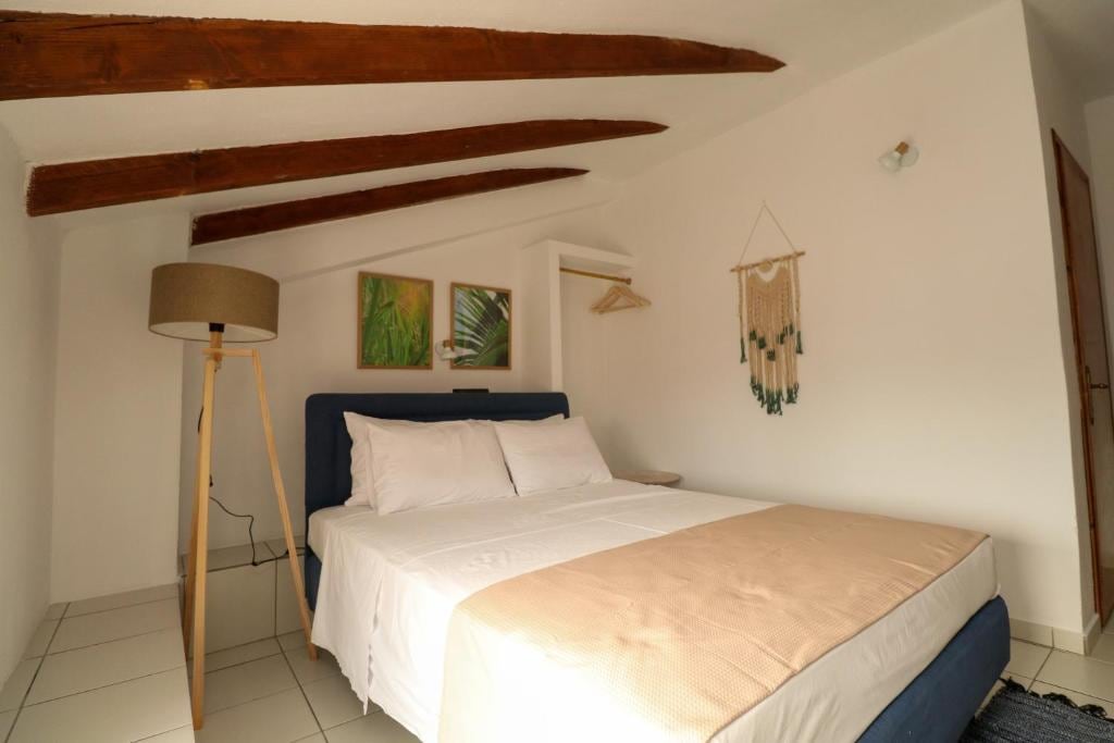 A bright sleeping area features a spacious bed with soft white linens and a neutral-toned blanket. Natural light fills the room through a window, and decorative elements include wall art depicting foliage and a macramé wall hanging. Exposed wooden beams enhance the room's character.
