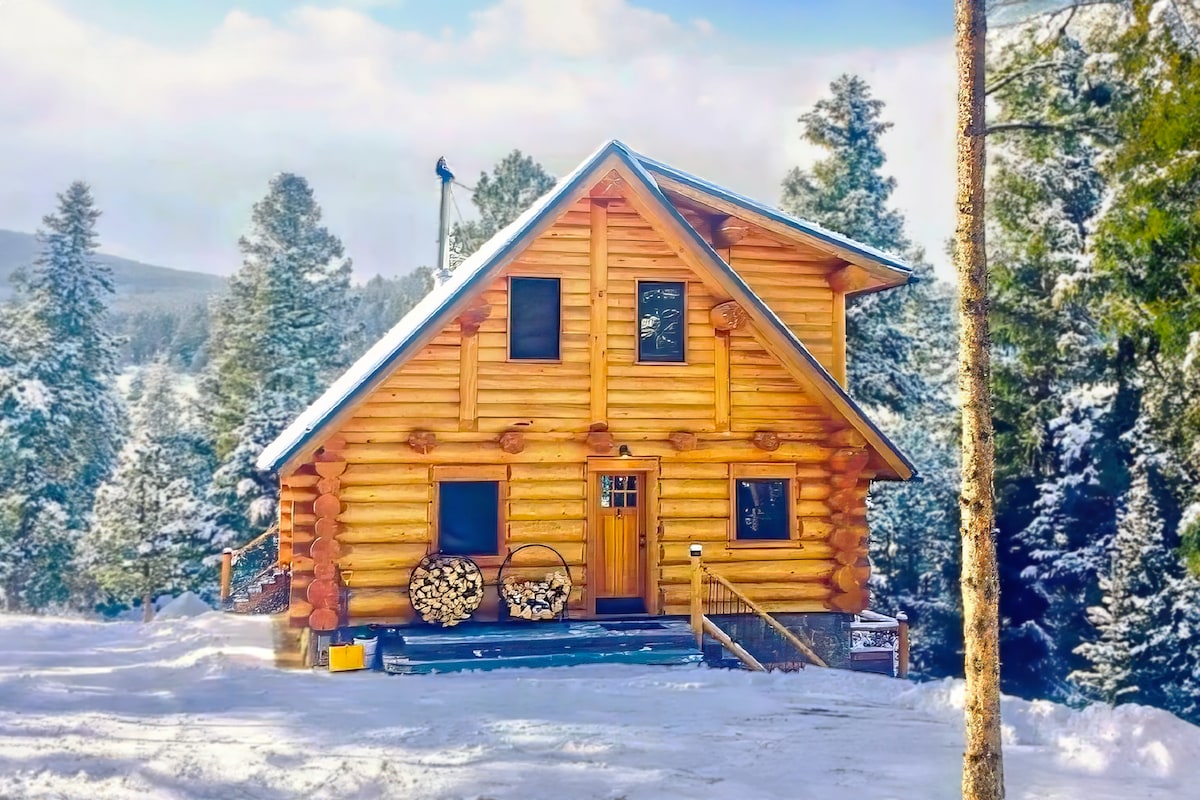 A handcrafted log cabin sits amidst a snowy landscape, framed by towering pine trees. The cabin features a welcoming front porch, a warm yellow door, and stacked firewood, providing a rustic charm. Snow blankets the surrounding area, enhancing the serene mountain setting.