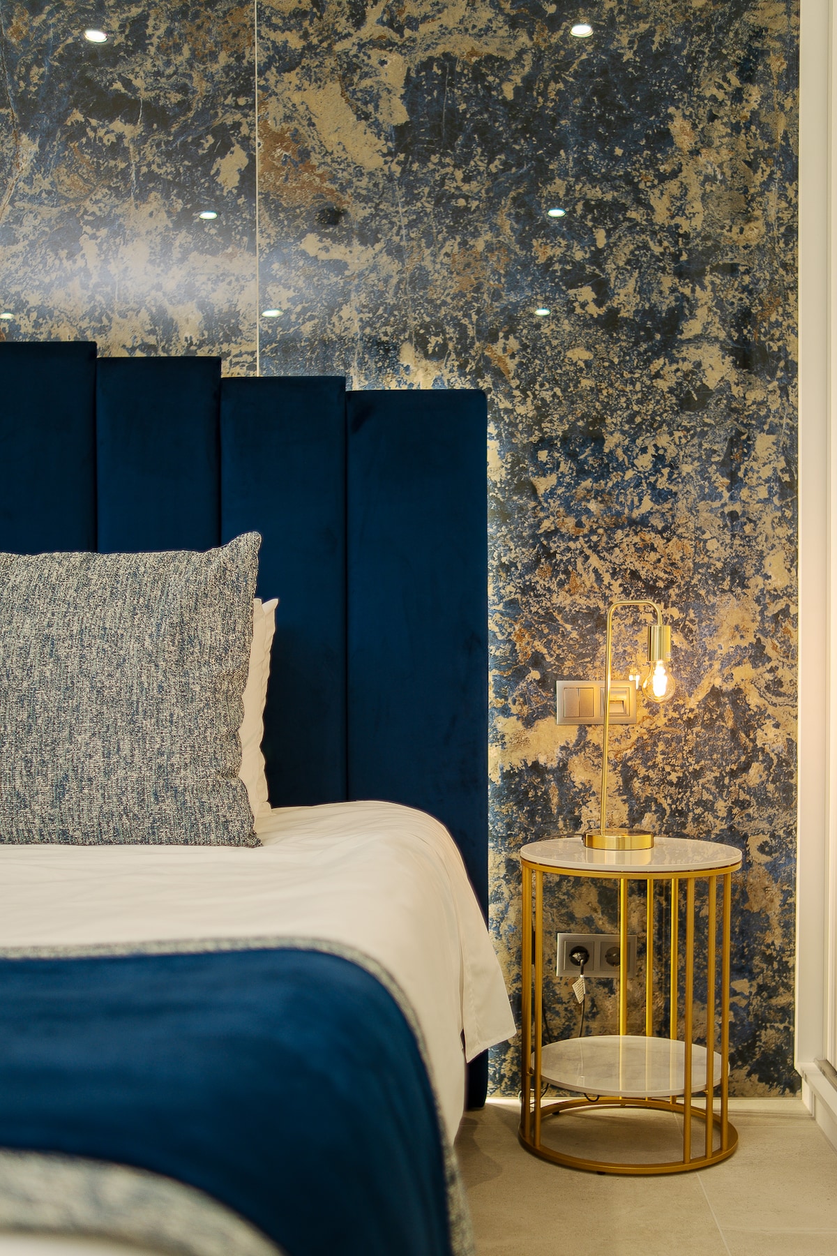 A spacious bedroom features a plush, navy upholstered headboard against a striking blue and gold marbled wall. A small, round side table with a marble top and a sleek golden base holds a stylish lamp providing soft lighting, enhancing the room's elegant design.
