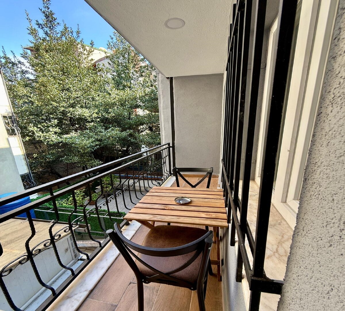 A private balcony is showcased with a wooden table and two chairs, offering a serene outdoor space. Lush green trees can be seen nearby, enhancing the tranquil environment. The railing features elegant black wrought iron detailing, providing both safety and aesthetic appeal.