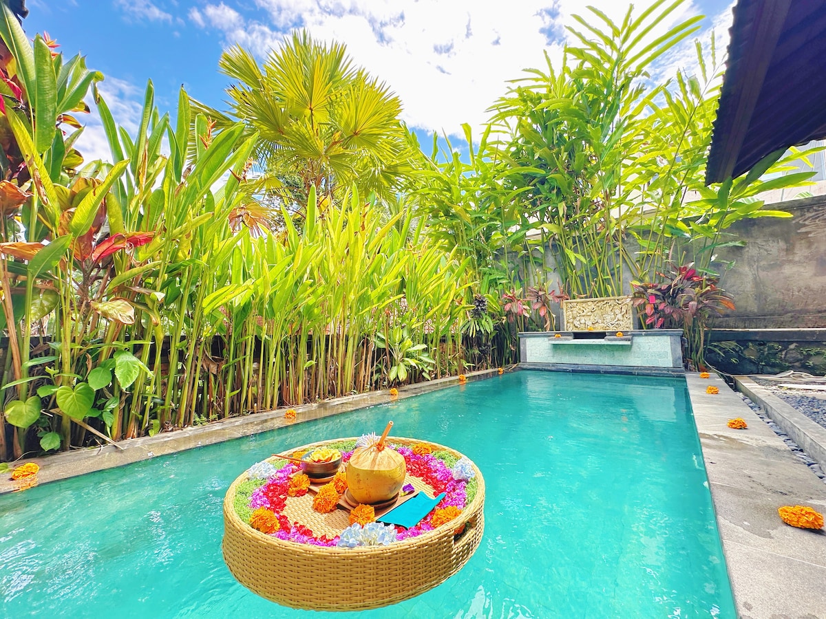A private pool surrounded by lush tropical greenery creates a serene setting. A decorative floating tray adorned with colorful flowers and traditional offerings is positioned in the water, enhancing the natural ambiance of this tranquil space.