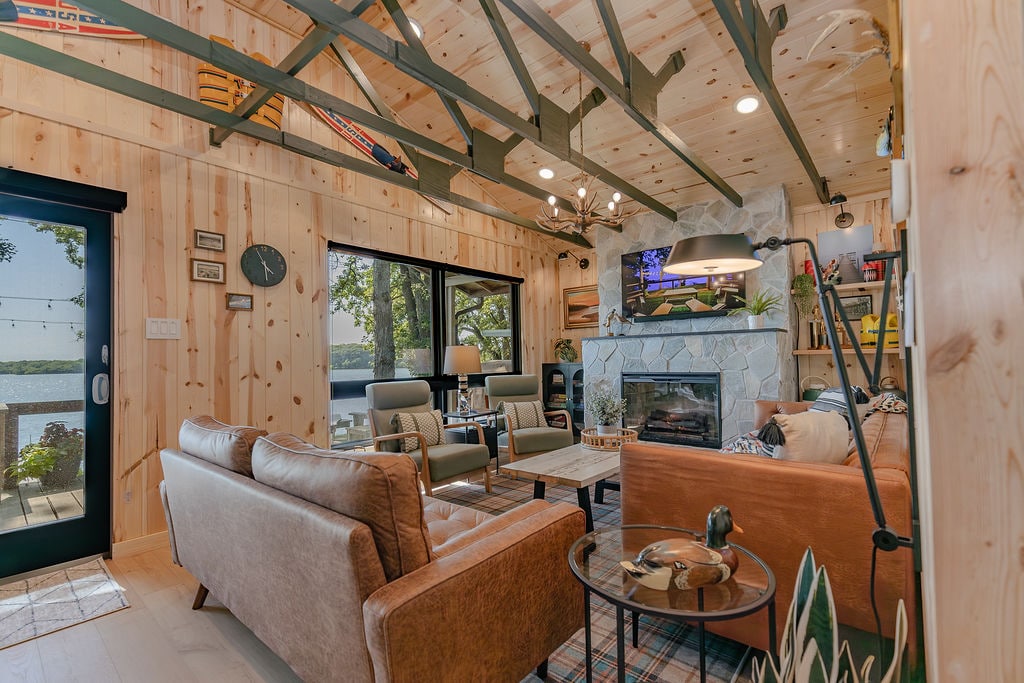 The living room features a blend of natural wood paneling and exposed beams, creating an airy environment. Relaxing sofas are positioned for lake views, while a smart TV is mounted above a modern electric fireplace. Large windows invite in ample natural light.