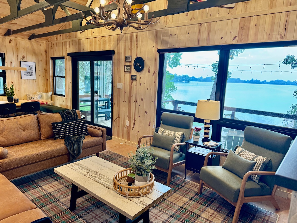 A spacious living room features large windows offering views of Little Spirit Lake. Comfortable sofas and armchairs are arranged around a centrally located coffee table. Natural wood paneled walls and a chandelier contribute to a cozy ambiance, with light filtering in to highlight the inviting decor.