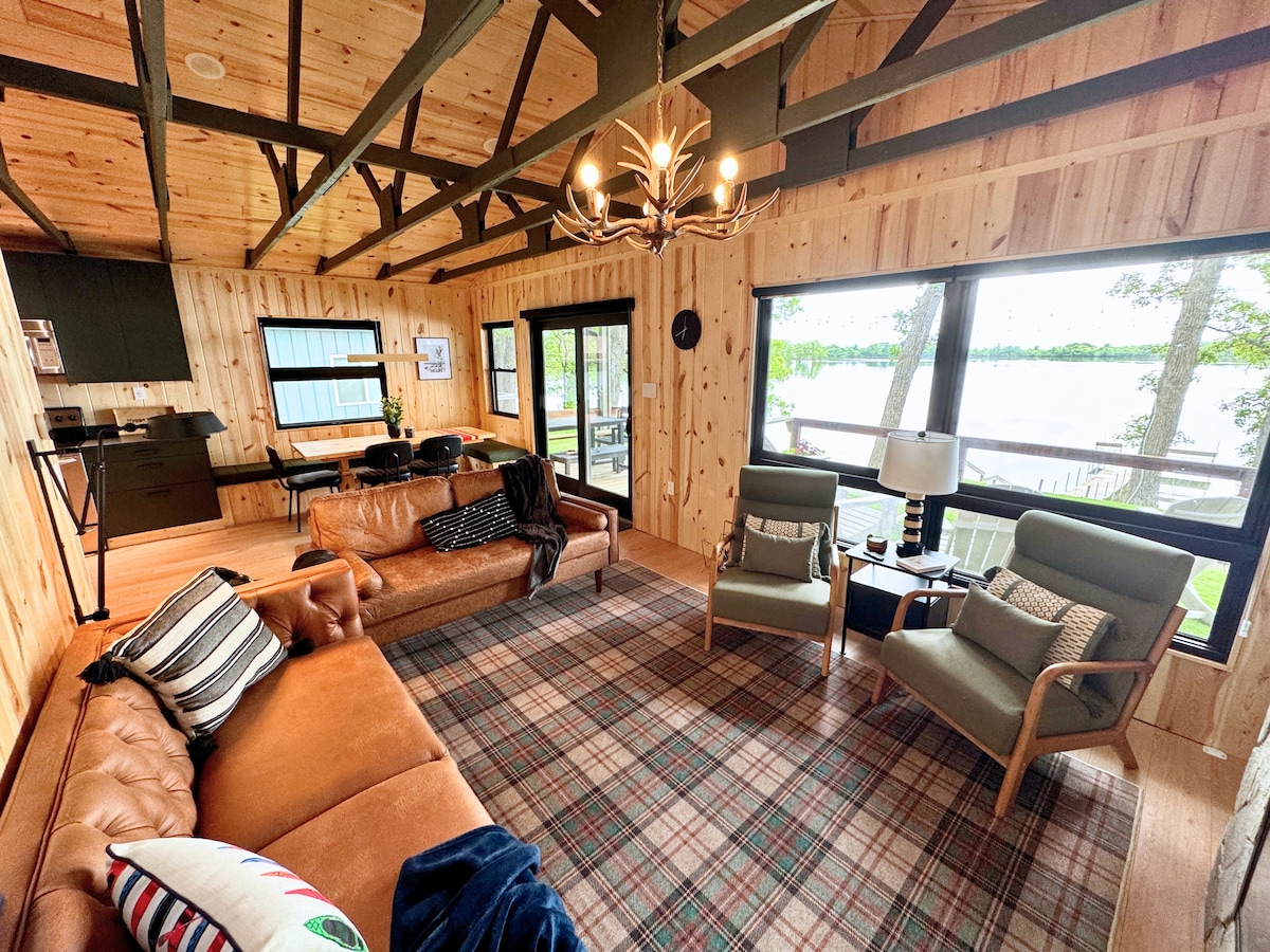 A spacious living area features wooden walls and beams, creating a cozy ambiance. A leather sofa, plaid rug, and armchairs offer comfortable seating. Large windows provide views of the outdoors, enhancing the connection to nature. A stylish chandelier adds a touch of elegance to the space.