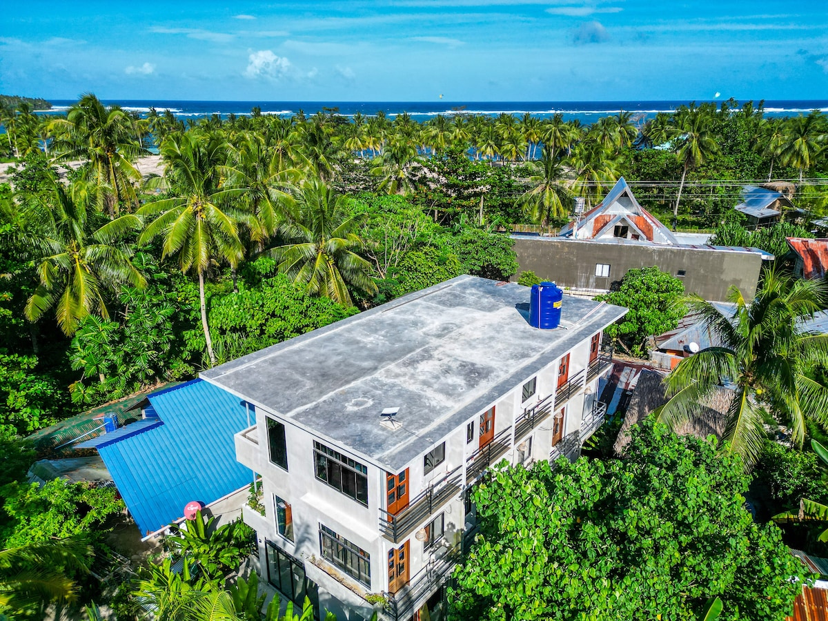An aerial view of a multi-story building is surrounded by lush greenery and palm trees. The ocean is visible in the background, and a blue swimming pool is located on the property. The roof features a water tank, indicating modern amenities.