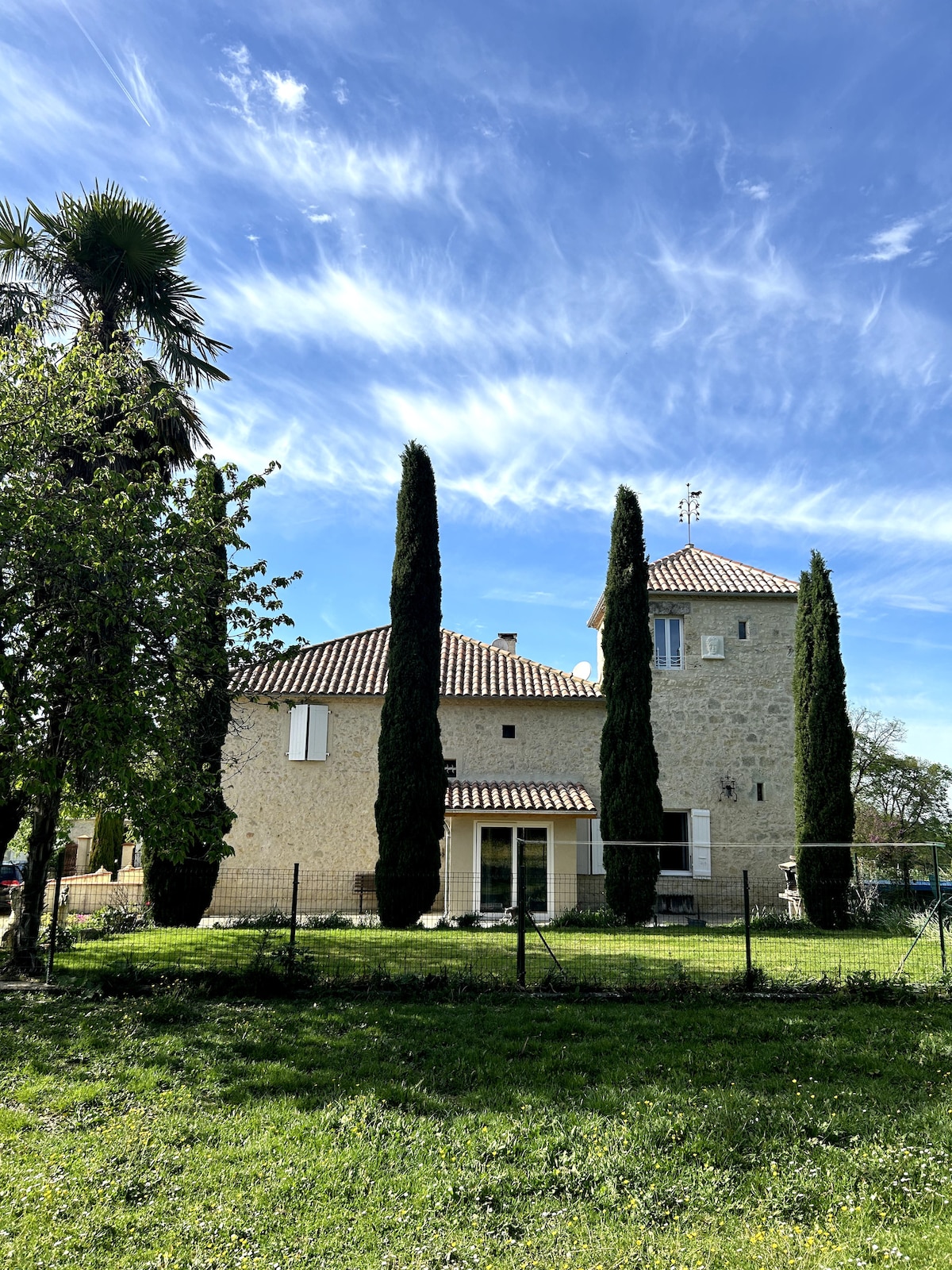 The exterior of the renovated stone house is framed by tall cypress trees and lush greenery. A charming terrace with a striped awning overlooks the garden. The roof features terracotta tiles and there are two white, shuttered windows visible. A peaceful atmosphere is suggested by the clear blue sky.