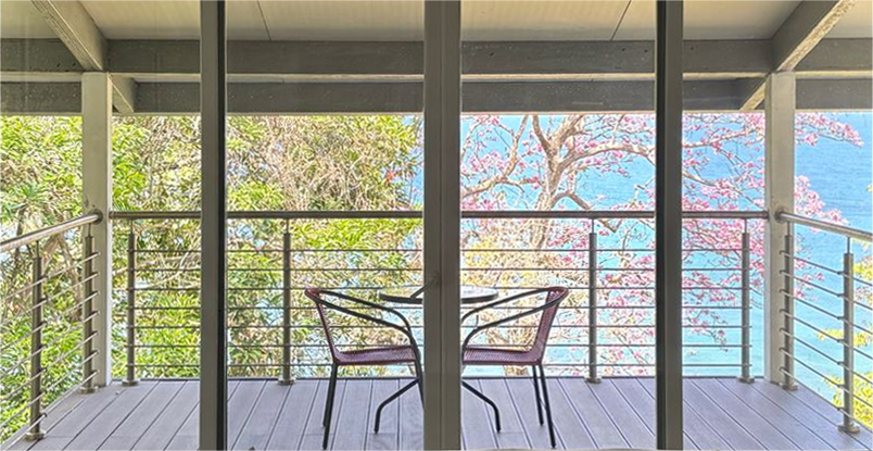 A serene balcony is viewed through glass doors, featuring a small table and two chairs. Lush trees and a vibrant pink flowering tree frame the backdrop, with a glimpse of the ocean visible in the distance, creating a tranquil outdoor setting.
