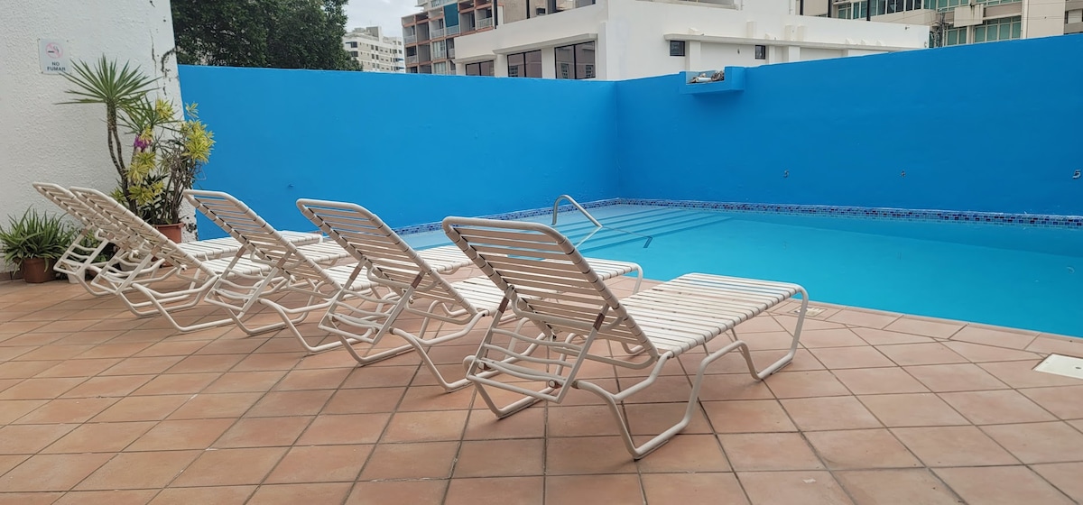 A pool area is depicted, featuring a clear blue pool surrounded by tiled flooring. Several white lounge chairs are arranged beside the pool, offering a spot for relaxation. Lush plants add a touch of greenery against the vibrant blue wall in the background.