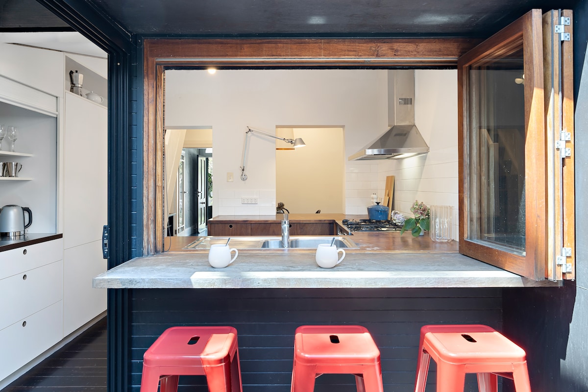 An open-air kitchen counter is featured, with three red stools positioned in front. A window allows for easy access between the indoor kitchen and outdoor space, illuminated by natural light. The kitchen includes a modern design with stainless steel appliances visible in the background.