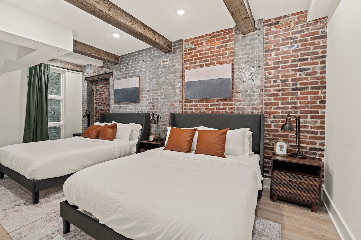 This spacious room features two beds with light bedding and contrasting orange accent pillows. Exposed brick walls add texture, complemented by wooden beams in the ceiling. Soft area rugs and nightstands with lamps complete the inviting atmosphere.
