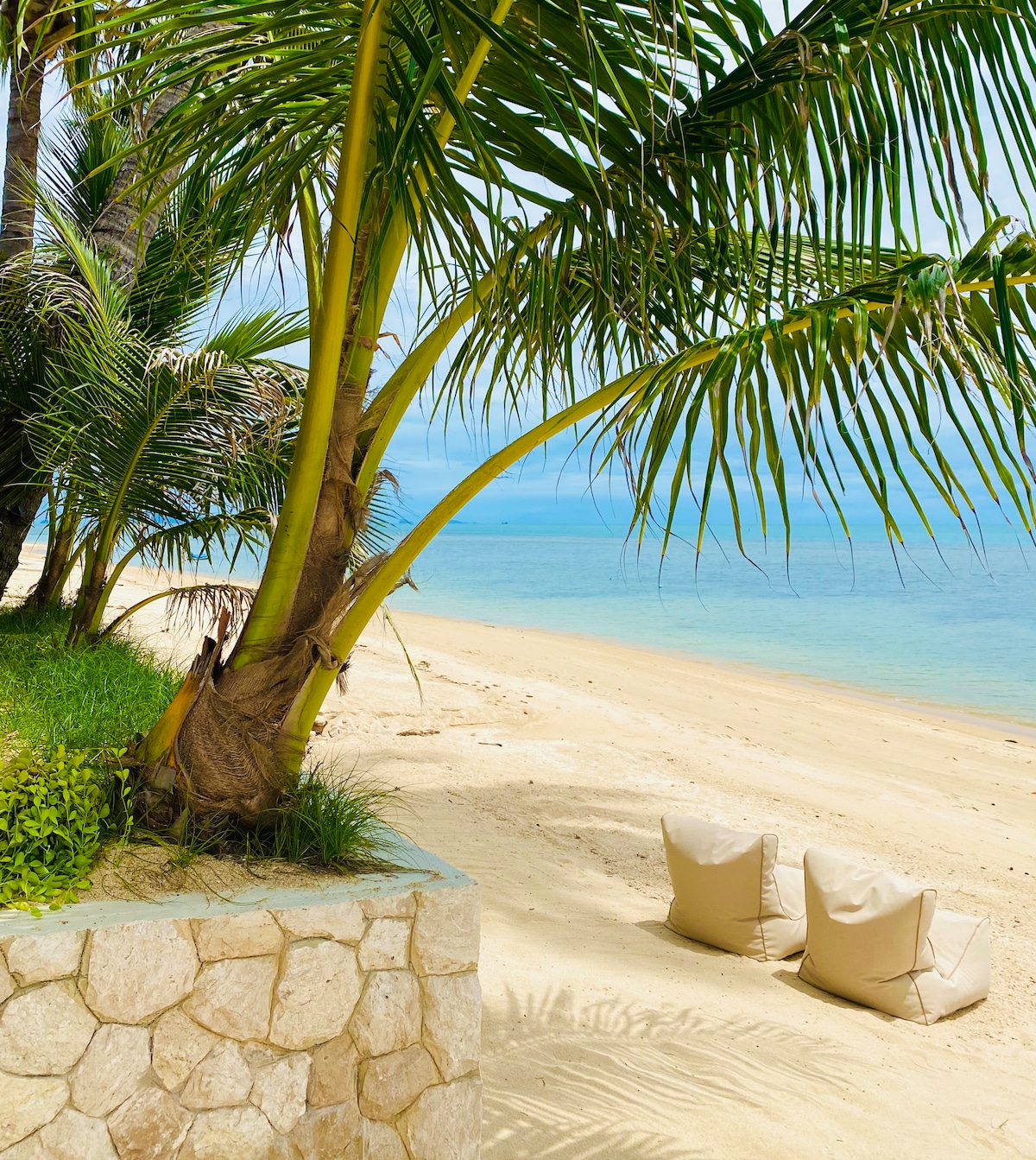 Two cushioned lounge chairs are positioned on a sandy beach, framed by palm trees. The calm turquoise water extends into the distance, complementing the tranquil atmosphere. A stone wall with lush greenery is visible in the foreground, enhancing the natural beauty of the setting.