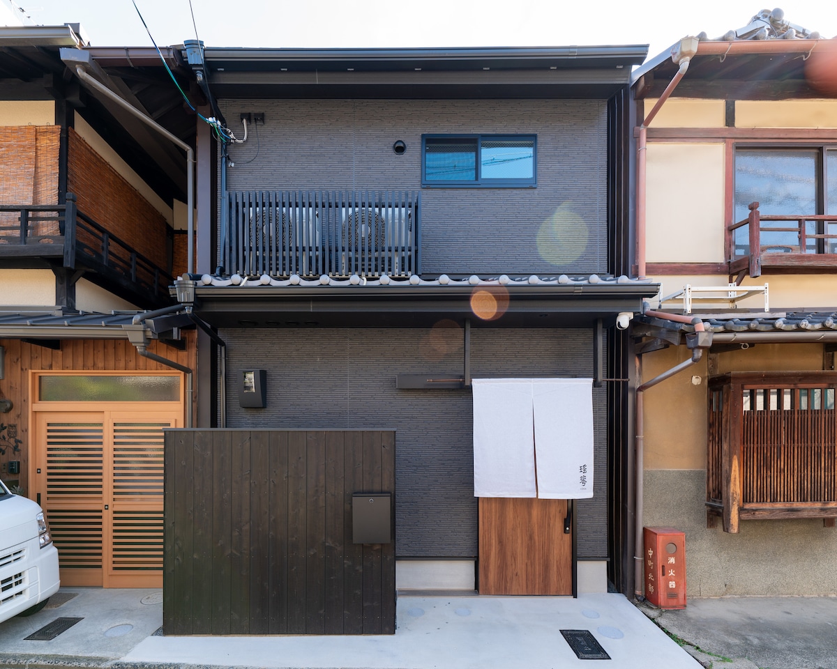 The exterior of a centuries-old house showcases a modern renovation with a dark façade, complemented by wooden accents. A white curtain hangs at the entrance, providing a subtle touch, while neighboring traditional wooden homes can be seen, highlighting the blend of old and new.