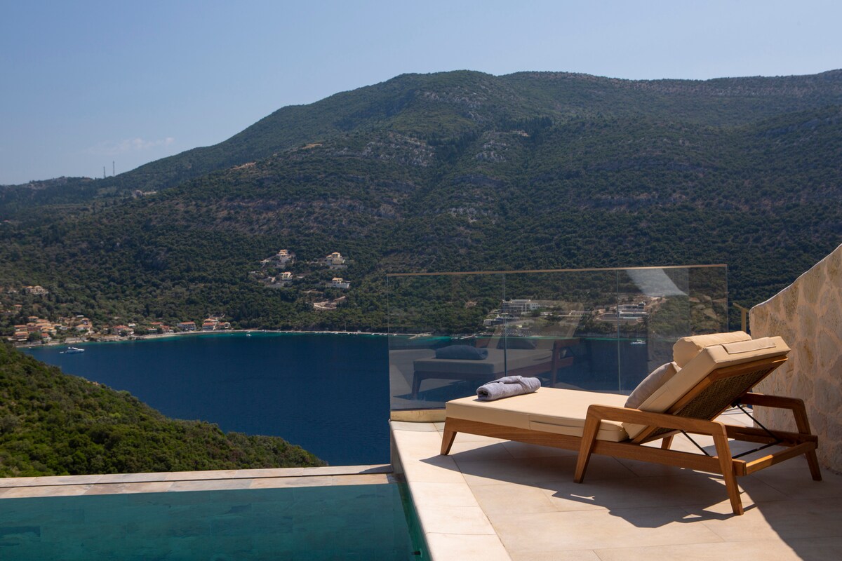 A sun lounger rests on a stone terrace overlooking a tranquil bay surrounded by lush mountains. The clear water reflects the bright sky, while the horizon showcases a peaceful coastal village, adding to the serene atmosphere of the villa's setting.