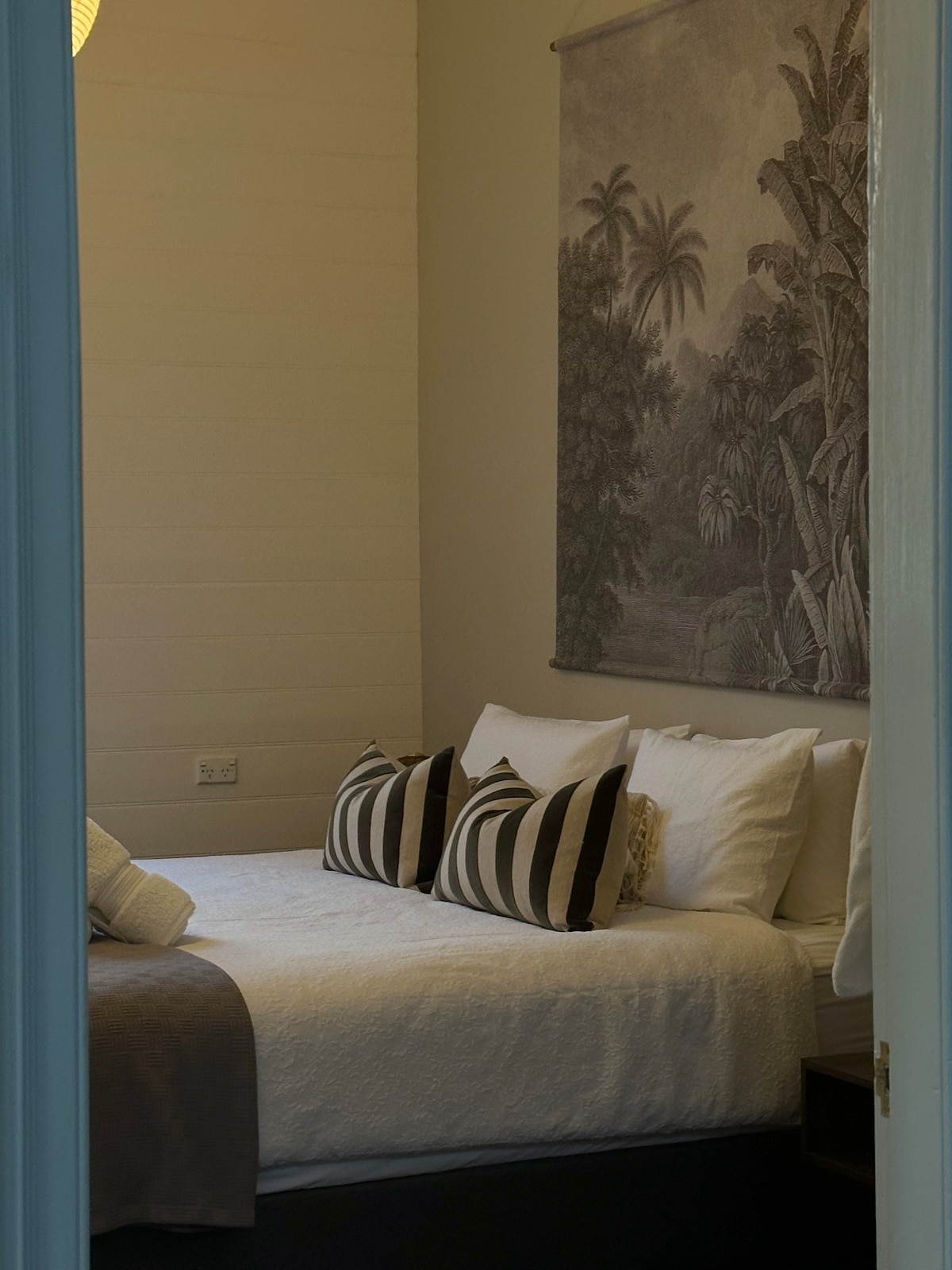 A compact sleeping area is visible, featuring a spacious bed with white linens and striped pillows. A decorative wall hanging adds a natural touch, while soft lighting enhances the calm atmosphere. The room is framed by an open doorway, providing glimpses of its tranquil setting.