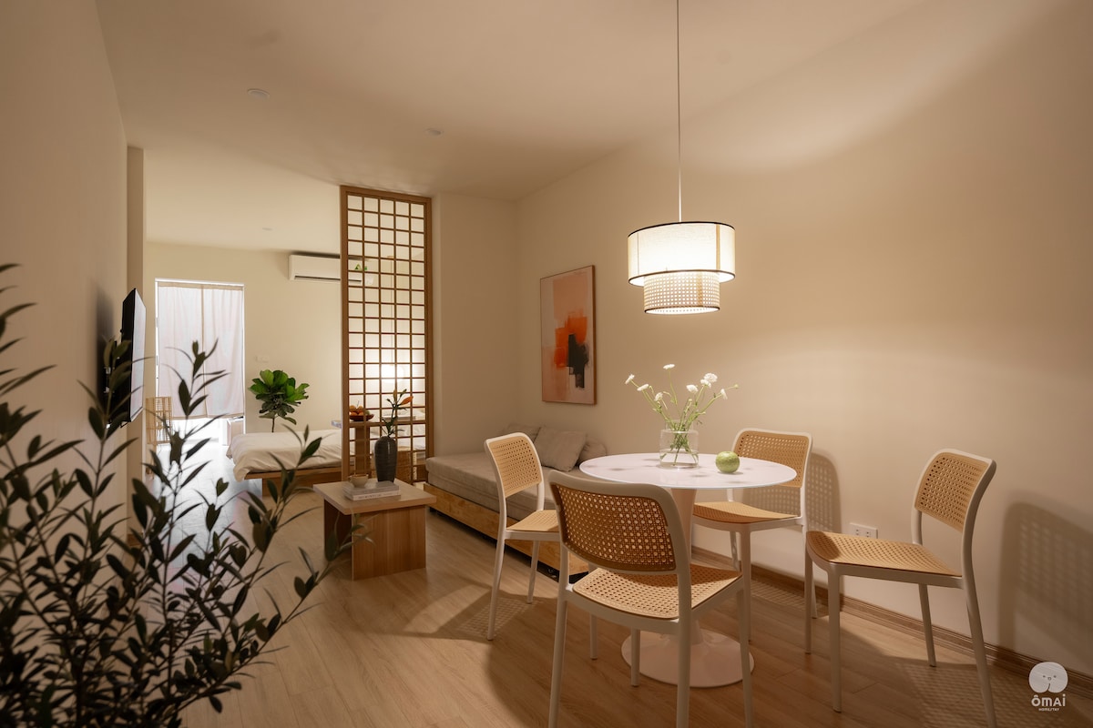 A dining area features a round table surrounded by four light-colored chairs. A simple pendant light hangs above, illuminating the space. Soft textures and plants bring subtle warmth, while a sliding screen divides the area from a cozy sleeping nook in the background.