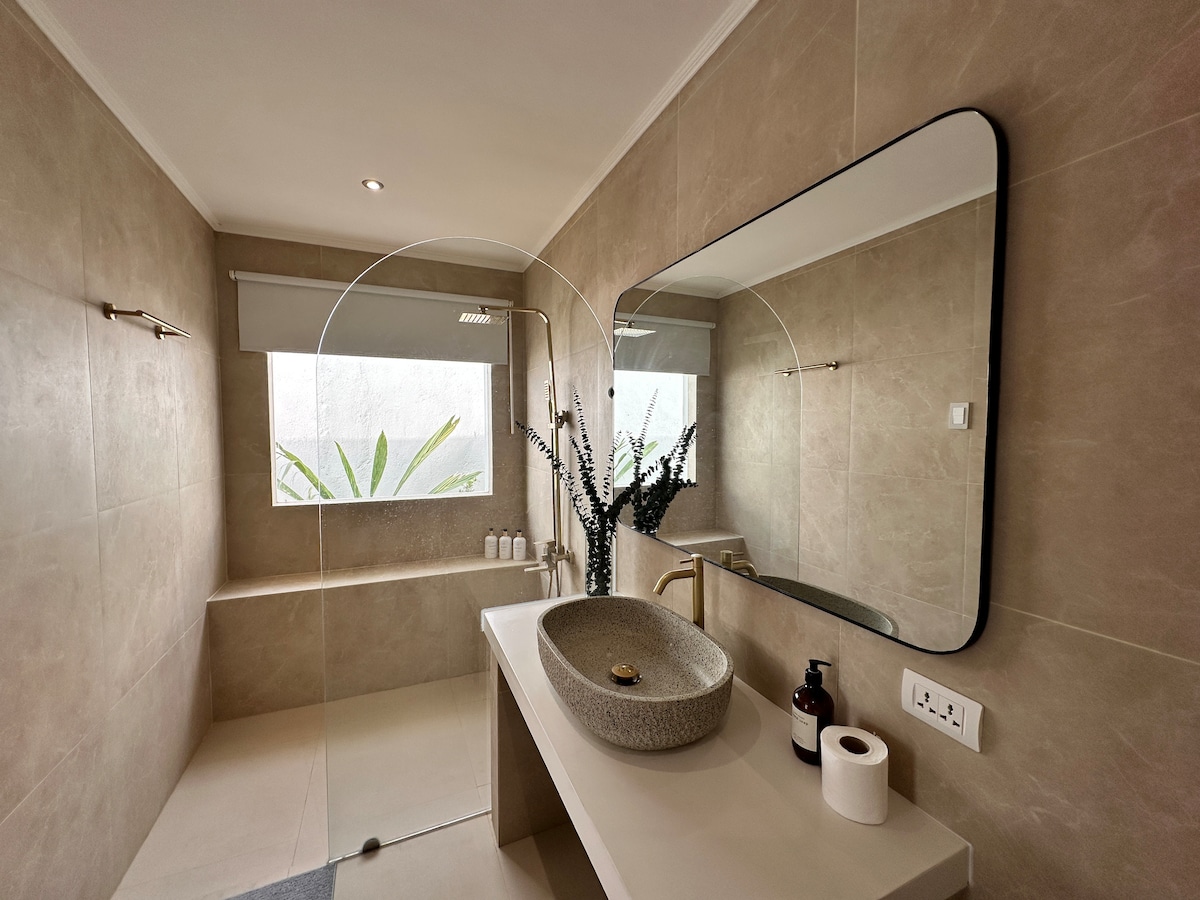 A modern bathroom features neutral-toned walls and a spacious layout. Natural light filters through two windows, illuminating a chic wash basin with a curved mirror above. Elegant toiletries are arranged on the countertop, with a touch of greenery visible outside through the glass.