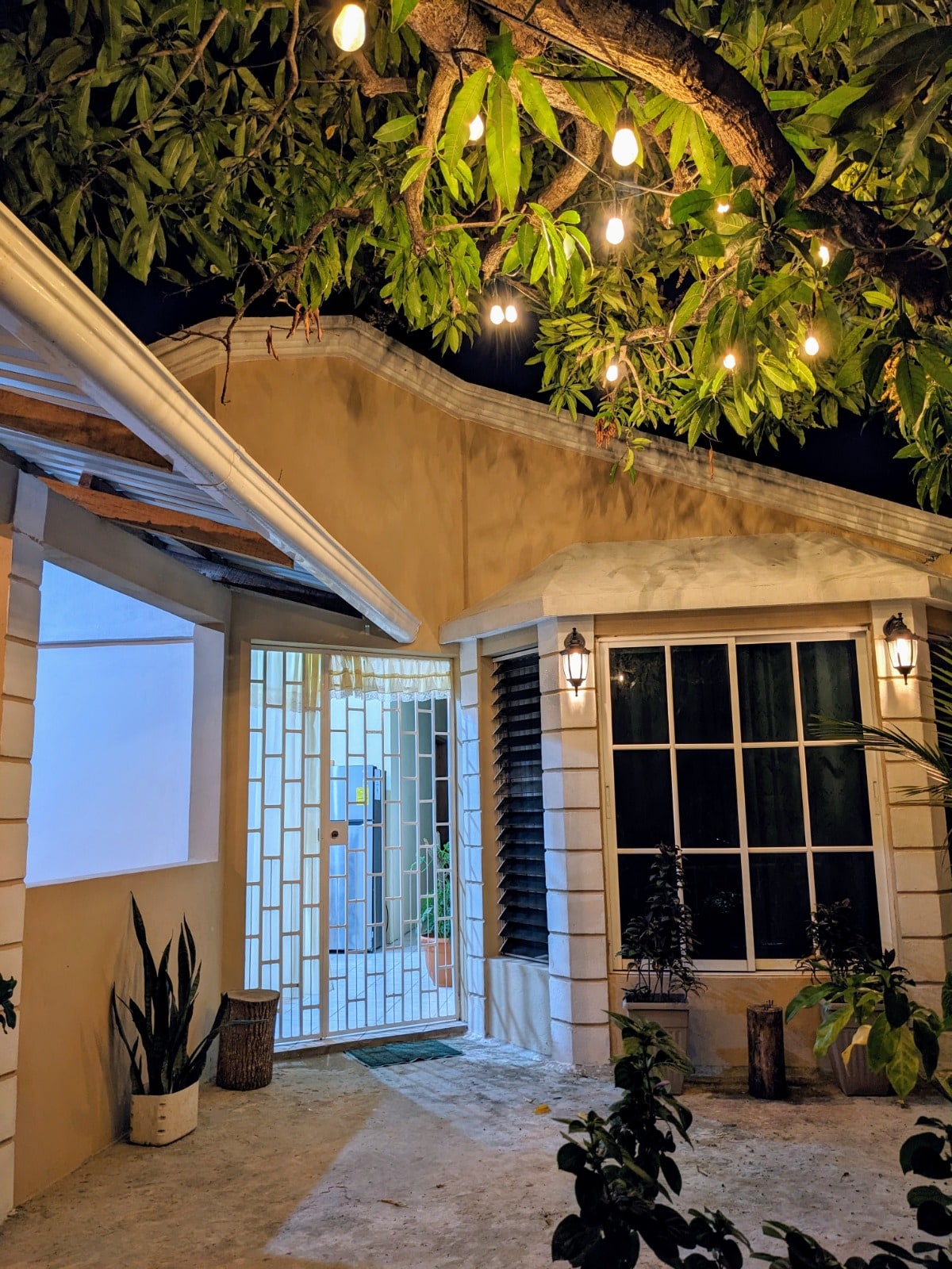 The entrance of the apartment is illuminated by soft lights hanging from a tree overhead. A door with a decorative grid invites entry, while a large window beside it offers a view of the outdoors. Lush greenery enhances the welcoming atmosphere.