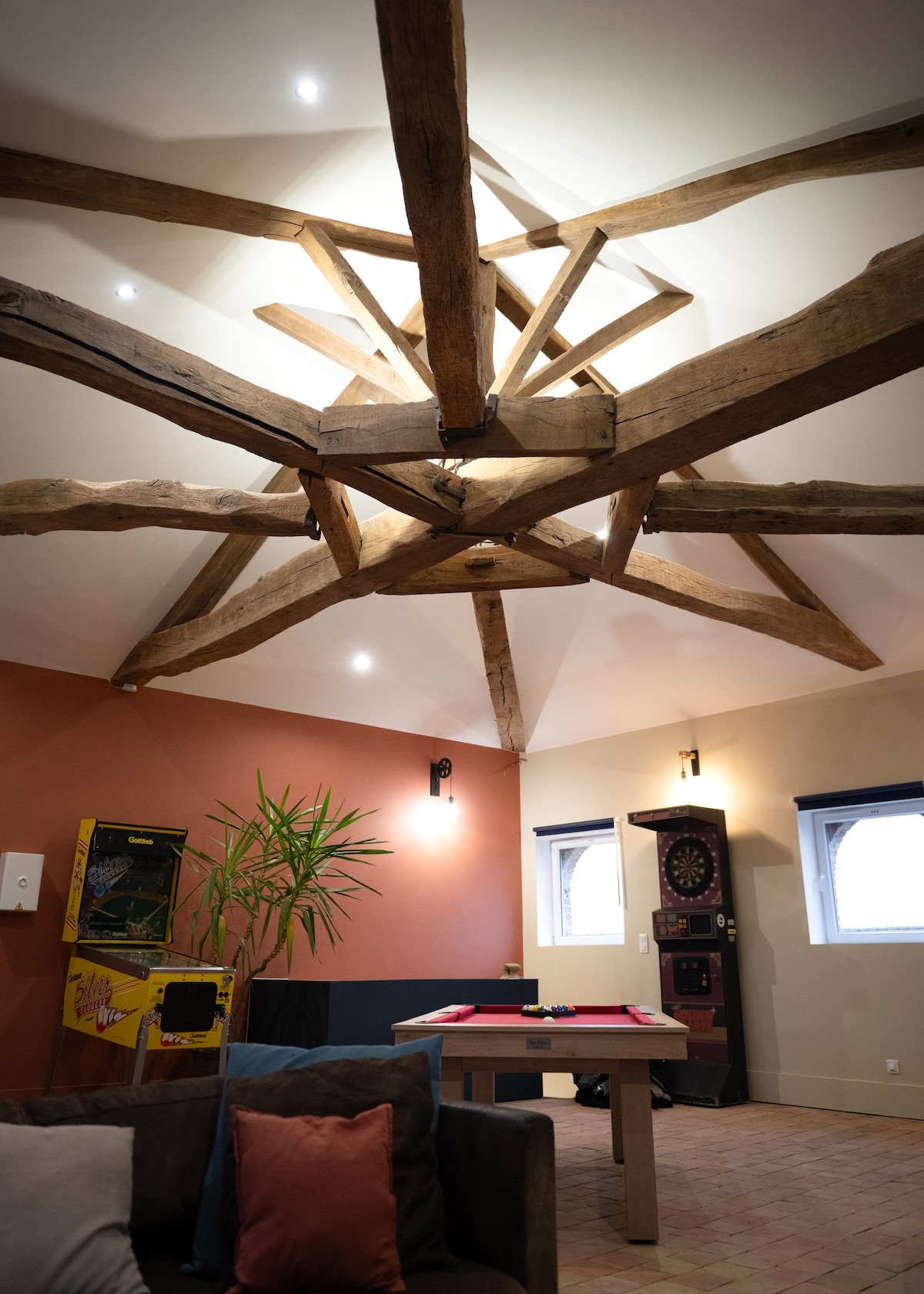 A spacious game room features a high ceiling with prominent wooden beams arranged in a star shape. One corner includes a dartboard and a vintage slot machine, while a large table provides space for games. Natural light streams through multiple windows, enhancing the inviting ambiance.