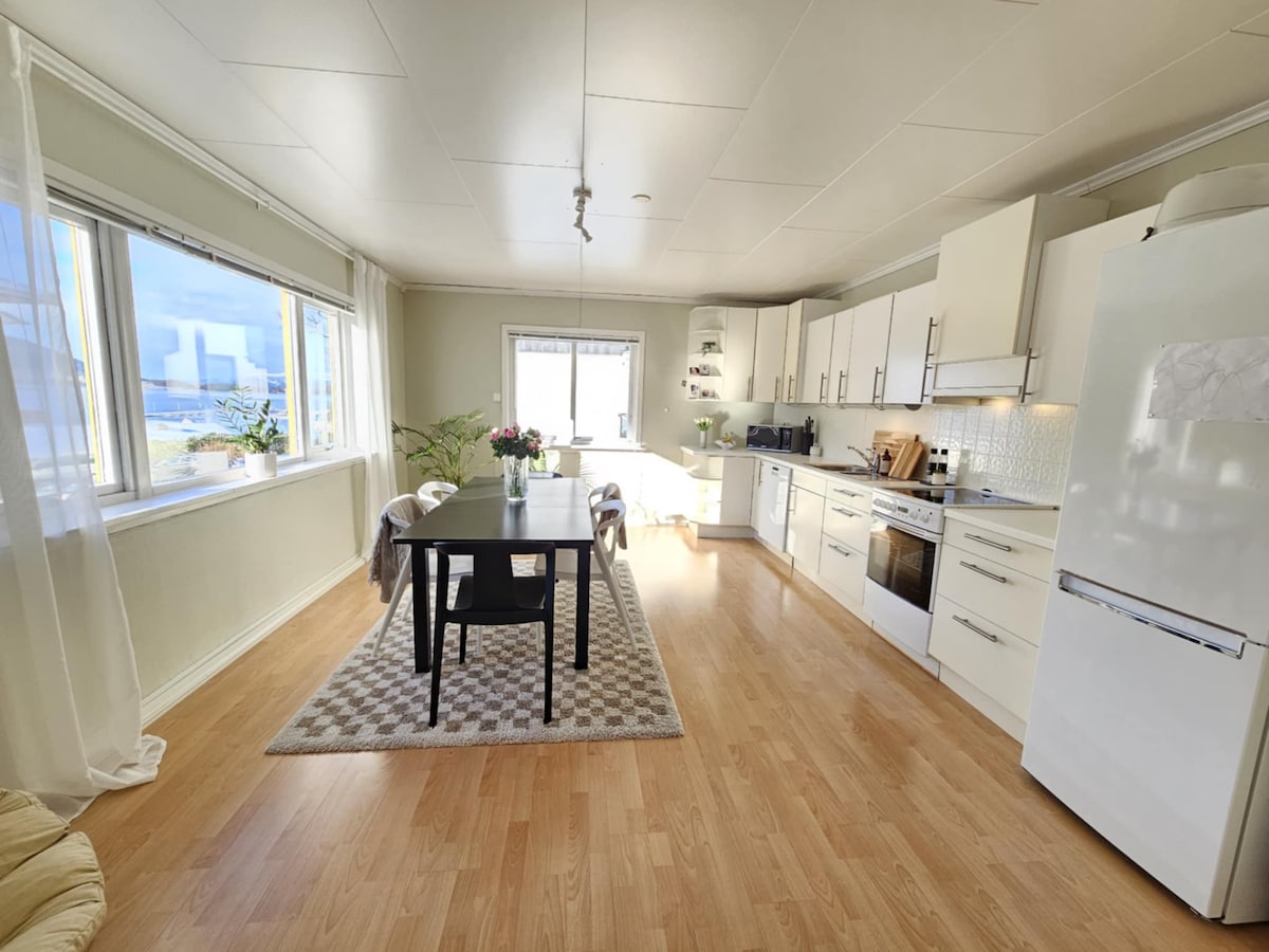 A spacious kitchen and dining area feature large windows allowing natural light to fill the room. A dark wooden table is surrounded by two chairs, complemented by a soft area rug. Modern kitchen appliances and cabinetry are neatly arranged along one wall.