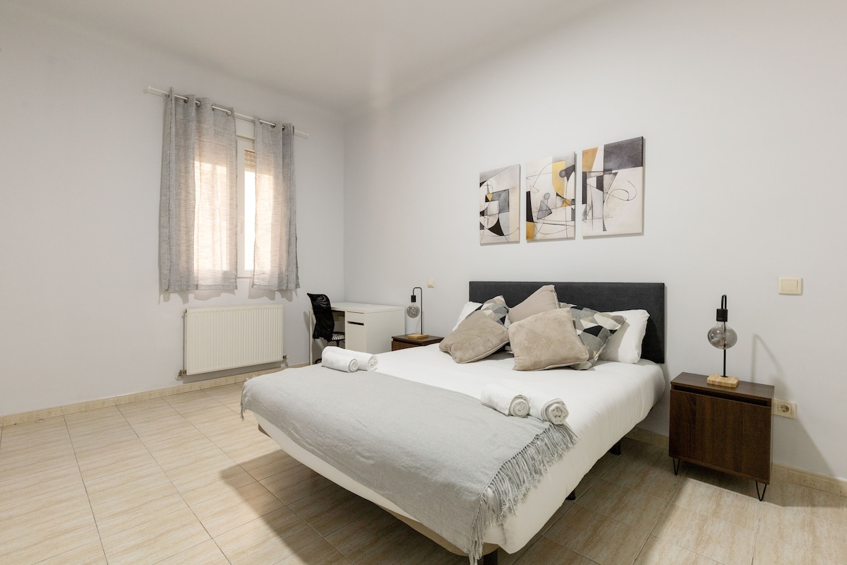 A well-lit bedroom features a spacious bed adorned with soft pillows and a light blanket. Two nightstands with lamps are positioned on either side of the bed. A decorative wall art piece enhances the room, while a window allows natural light to enter, complemented by sheer curtains.