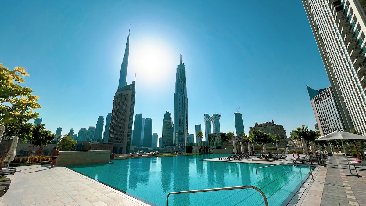 A gleaming rooftop pool features clear blue water, reflecting the sunlight. Surrounding the pool are lounge chairs and umbrellas, with views of the iconic Burj Khalifa and the Dubai skyline in the distance. Tall buildings and palm trees frame the scene, enhancing a sense of tranquility.