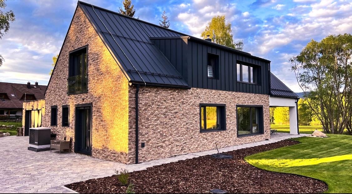 The exterior of a modern house is portrayed, featuring a mix of stone and dark panels. Large windows allow natural light to enrich the interior. A stone pathway leads to a landscaped yard, and a seating area is visible at the entrance, surrounded by well-maintained grass.