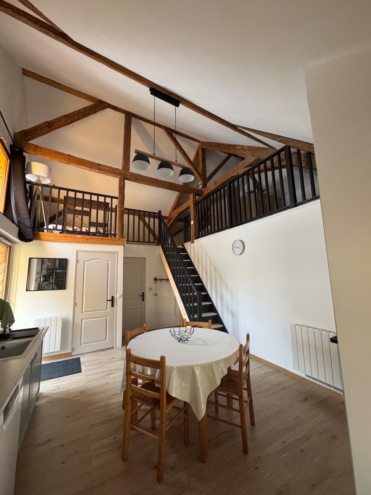 An open space loft design features high ceilings with exposed wooden beams. A circular dining table with wooden chairs is positioned on the floor, while a staircase leads to a mezzanine area. A clock is mounted on the wall, and a window allows natural light to enter.