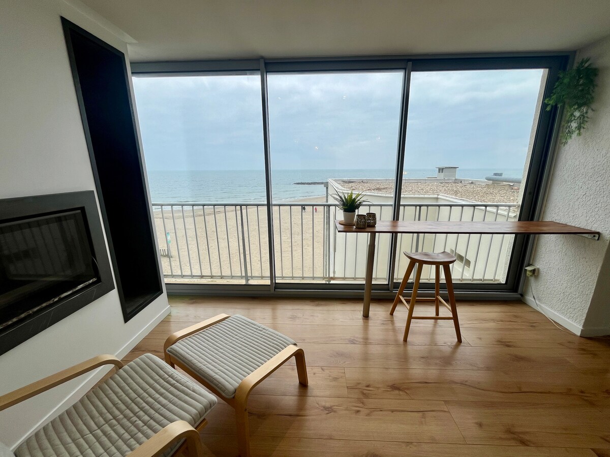 A cozy seating area is positioned near large glass windows, offering unobstructed views of the beach and sea. Two wooden chairs are placed beside a narrow table, creating a serene spot to relax while gazing at the tranquil water.