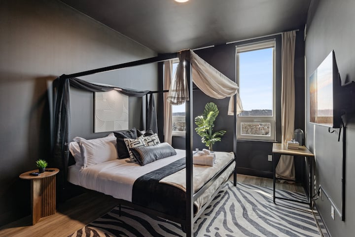 סוויטה ראשית עם קירות שחורים דרמטיים, מיטת קינג סייז עם חופה, שטיח זברה וחלונות גבוהים שמסביב נוף לעיר. הנכס שלנו להשכרה משלב עיצוב מודרני עם נוחות מפלט נעימה - מתאים למשפחות לשהייה ב-Airbnb ומושלם למפגשים ולמנוחה.