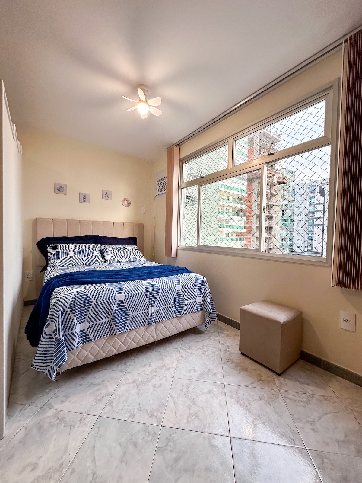 A bright bedroom is presented with large windows allowing natural light to fill the space. A spacious bed with a blue and white patterned blanket is centered against a light-colored wall. A small stool is positioned at the foot of the bed, adding functional decor.