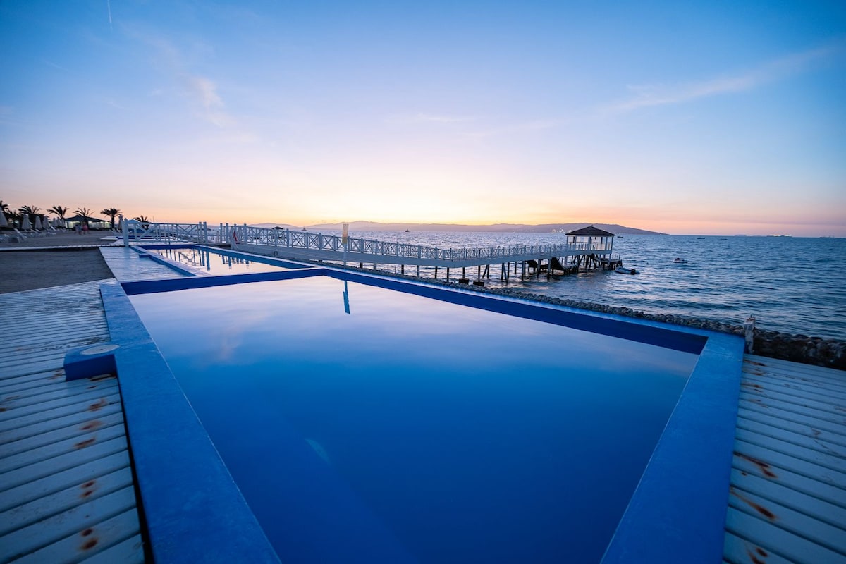 An infinity pool blends with the sea at sunset, showcasing a smooth transition between water and the horizon. A wooden pier extends into the water, with a gazebo visible at its end, complemented by the soft colors of the sky reflecting on the pool's surface.