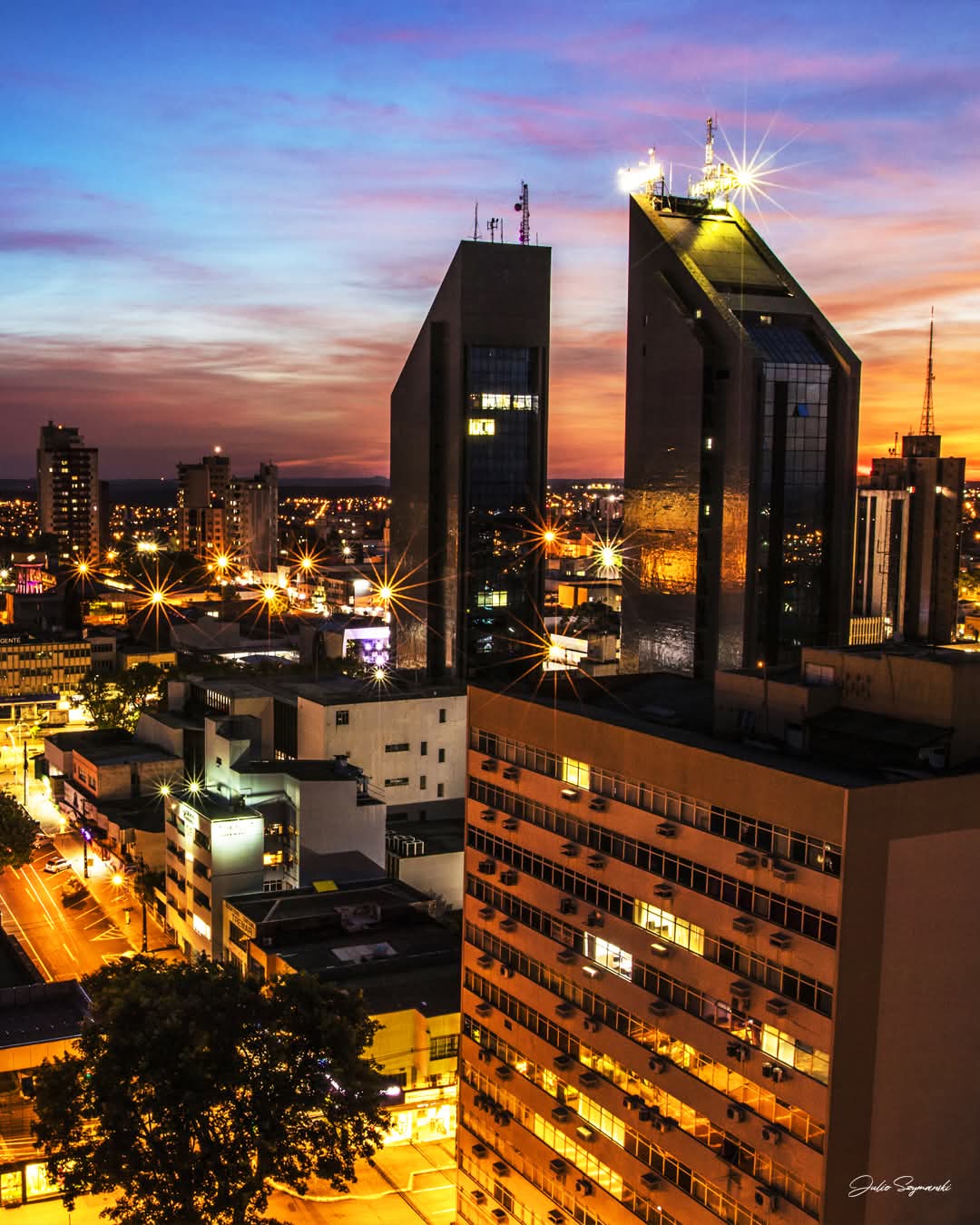 A panoramic view of a city skyline at dusk, highlighting modern buildings with reflective glass facades. Street lights twinkle below, illuminating roads that lead through the urban landscape. A colorful sky transitions from orange to deep blue, adding depth to the evening scene.