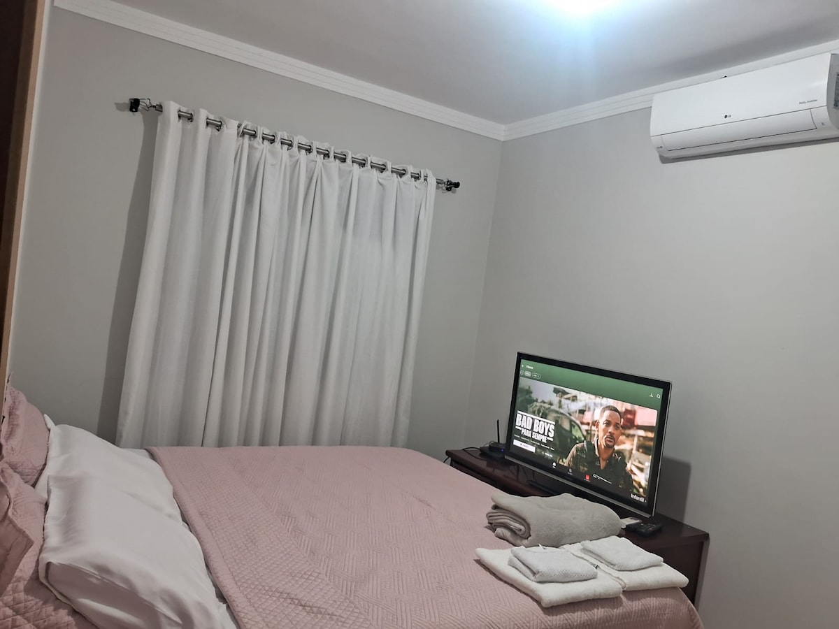 A well-appointed sleeping area features a spacious bed covered with a pink blanket. A television is positioned across from the bed. Soft, white curtains frame a window, while an air conditioning unit is mounted on the wall, providing comfort in a serene environment.