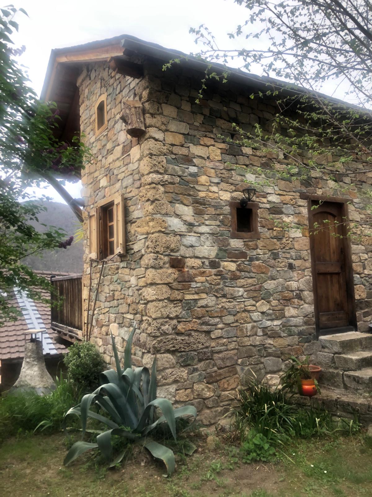 The exterior of a charming stone cottage is shown, showcasing a textured stone façade and wooden accents. Windows with shutters add to its character. A small staircase leads to the entrance, surrounded by greenery and plants, creating a serene and natural atmosphere.