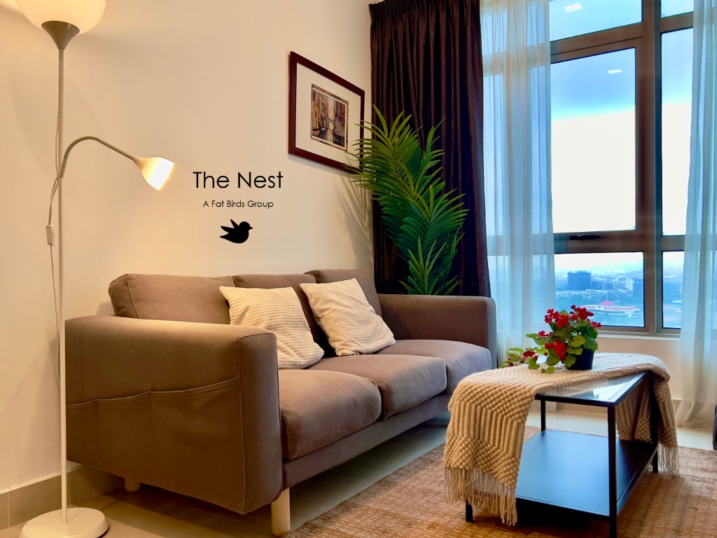 A modern living area features a comfortable grey sofa adorned with neutral throw pillows. A small table displays a vibrant plant and a decorative item. Natural light pours through large windows, framed by sheer and dark curtains, adding warmth to the space.