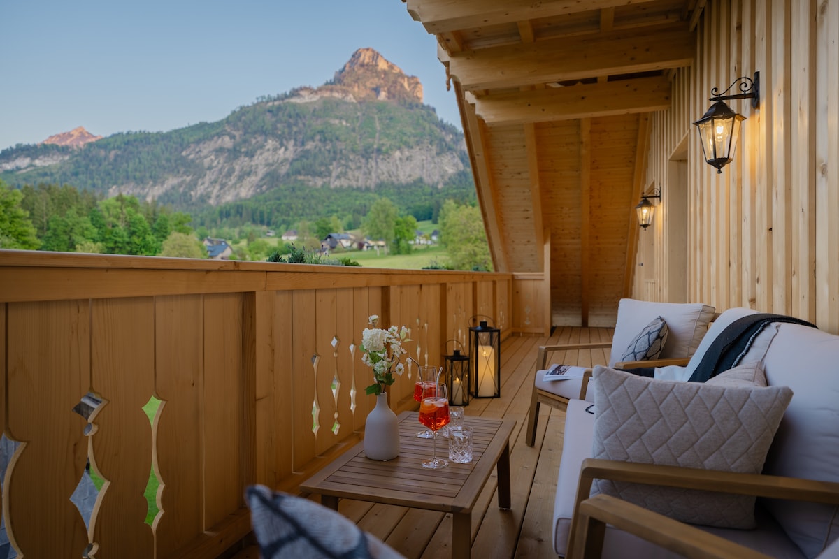 A charming balcony area features a wooden table and comfortable seating, adorned with decorative cushions. Lanterns provide soft lighting, while a scenic view of mountains is visible in the background. Drinks are placed on the table, enhancing the tranquil setting.