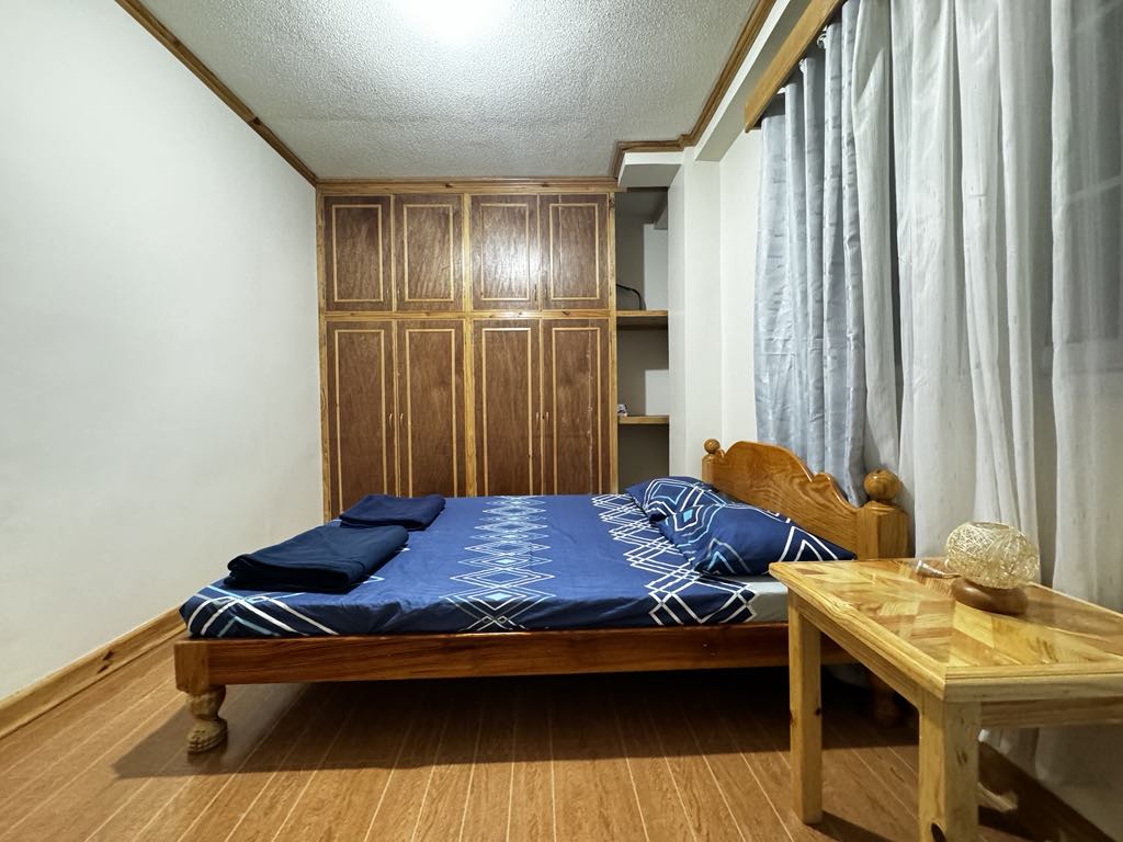 A cozy private room is showcased with a double-sized bed, adorned with blue bedding and additional blankets. A spacious wooden closet is positioned against the wall, alongside a small wooden table with a decorative item. Natural light filters through sheer curtains.