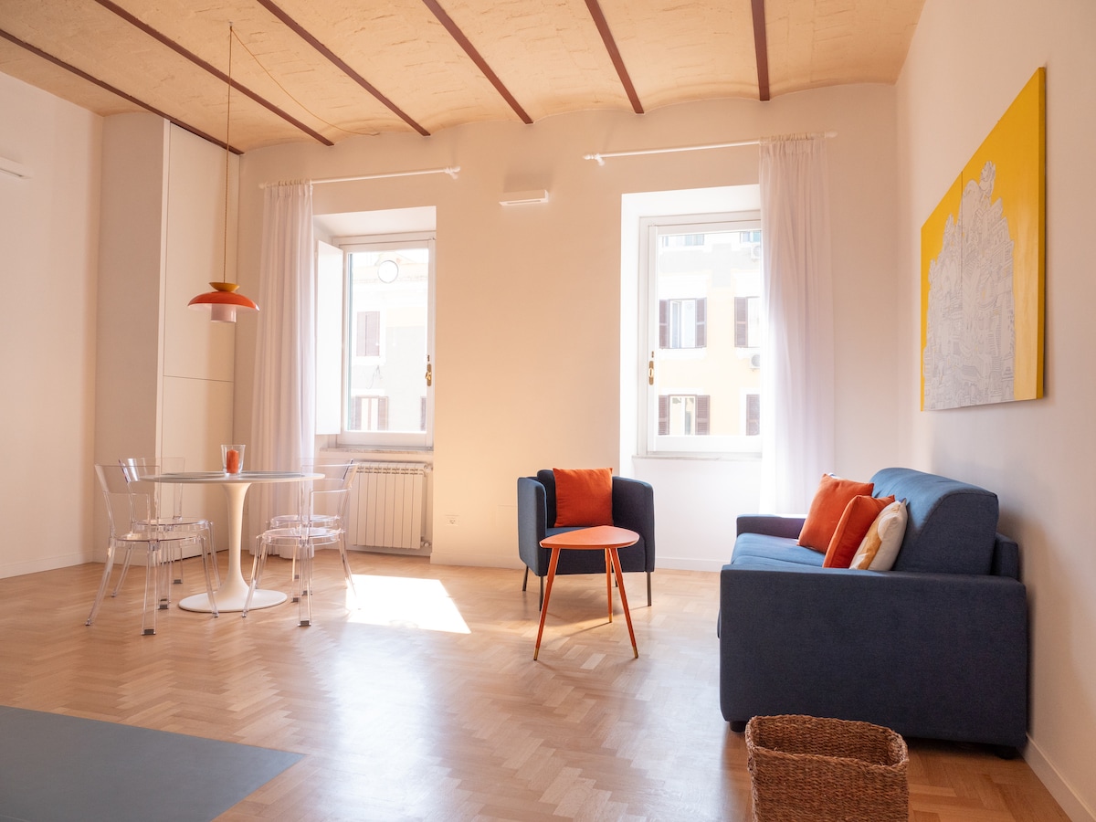 A bright living area features a comfortable blue sofa with contrasting orange cushions and a small round table set for two. Large windows allow for natural light to fill the space, while light wooden flooring adds warmth. An artwork hangs on the wall, adding a pop of color.