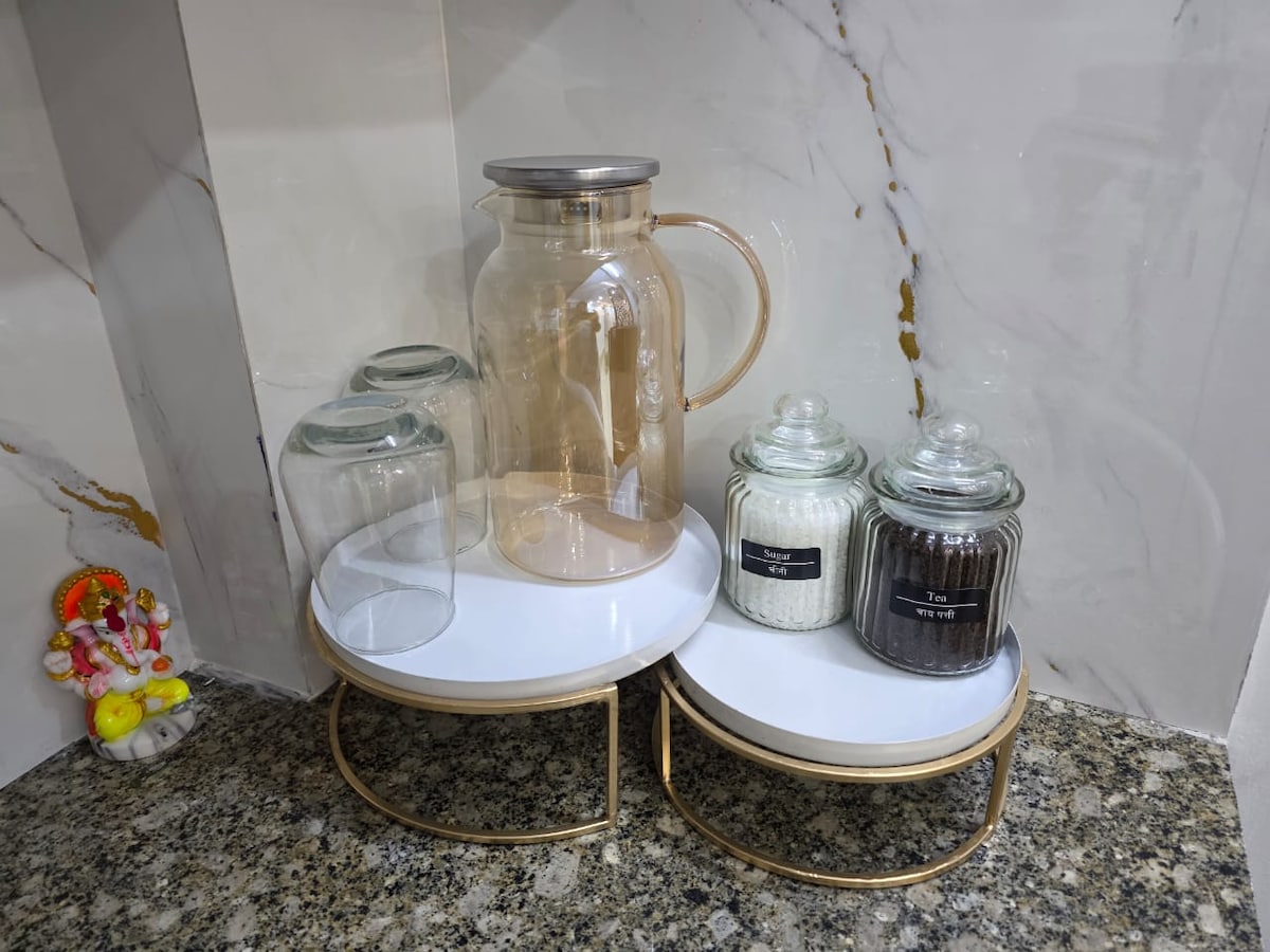 A kitchen countertop is shown featuring a glass pitcher and four glass containers. Two glass jars are labeled, containing sugar and tea, while the other jars are empty. The countertop has a smooth granite surface and a decorative figure is visible in the background.