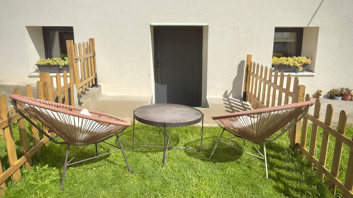 A small private garden area features two comfortable lounge chairs positioned around a circular table. The space is enclosed by a wooden fence, providing a cozy outdoor setting. A door leading to the studio is visible in the background, along with flower boxes adorning the windows.