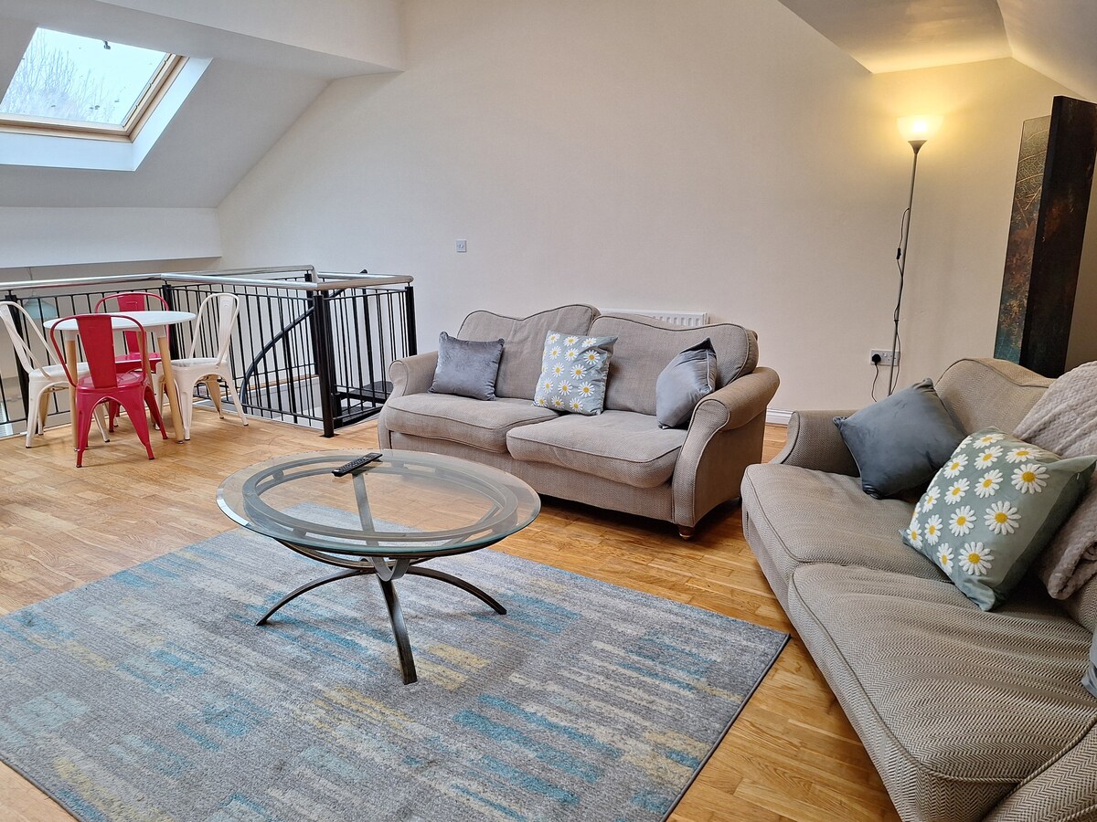 An open living area features two sofas arranged around a glass coffee table, with soft pillows for comfort. A patterned area rug lies beneath, complementing the warm wood flooring. Natural light fills the space through a skylight, enhancing the airy atmosphere.