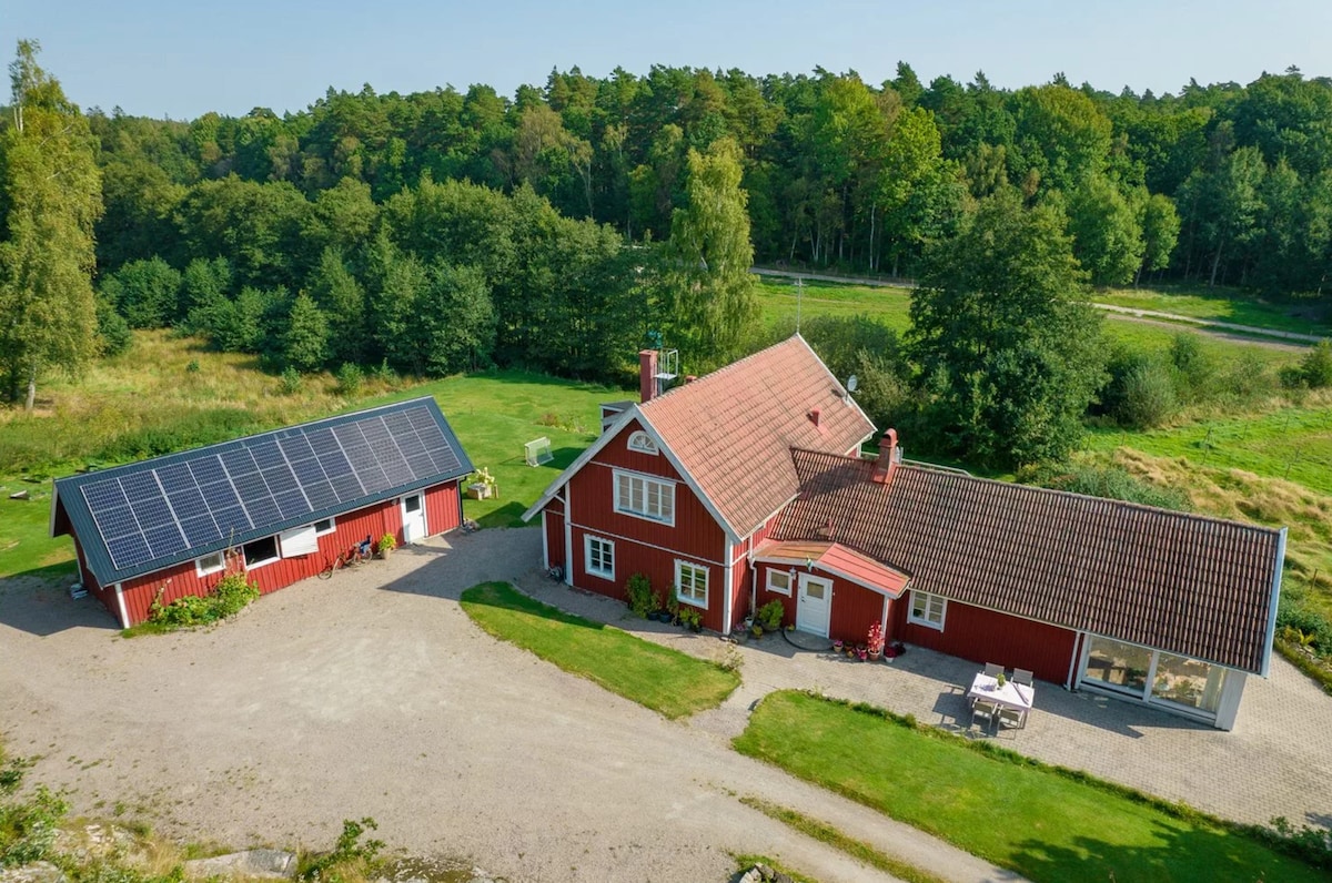 An aerial view captures the spacious red house and neighboring guesthouse, featuring solar panels on the roof. Lush greenery and trees surround the property, emphasizing its secluded location. A gravel pathway leads to the entrance, inviting gatherings in a natural setting.