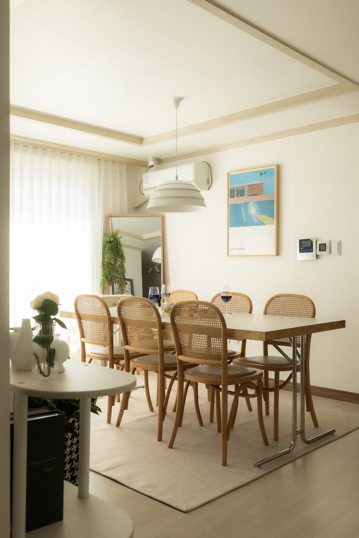 A dining area features a light wooden table surrounded by six rattan chairs. Natural light streams in through sheer curtains, illuminating the space. A wall-mounted mirror reflects the ambiance, complemented by a contemporary piece of artwork above the table.